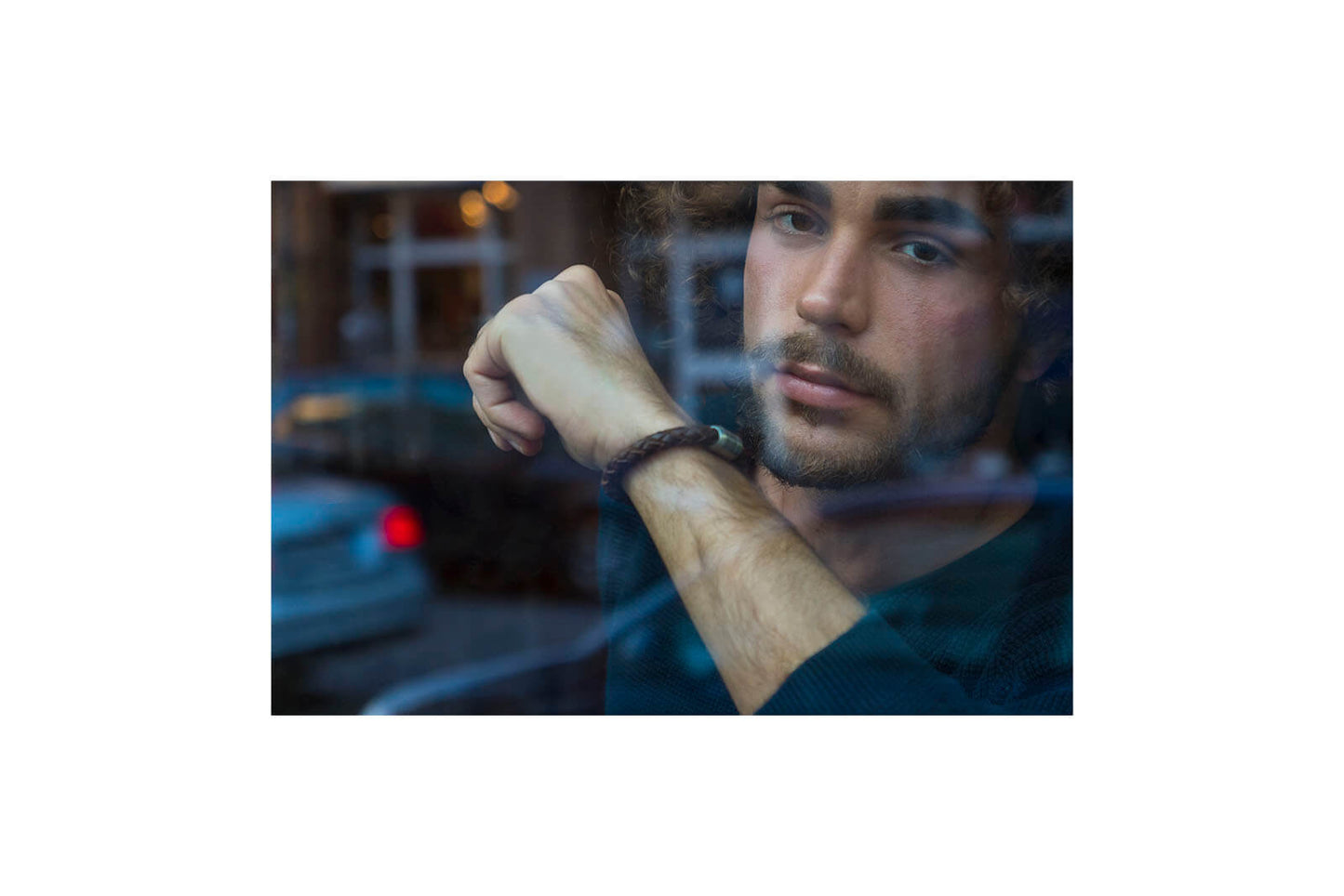 Junger Mann schaut aus Cafe heraus und zeigt sein geflochtenes, braunes Lederarmband mit Edelstahl Gravur