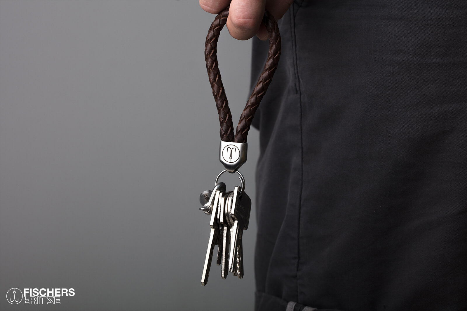 Hand haelt braunen Schluesselanhaenger Leder geflochten mit Schluesselbund, Edelstahlfassung mit Gravur sichtbar