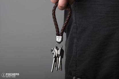 Hand haelt braunen Schluesselanhaenger Leder geflochten mit Schluesselbund, Edelstahlfassung mit Gravur sichtbar