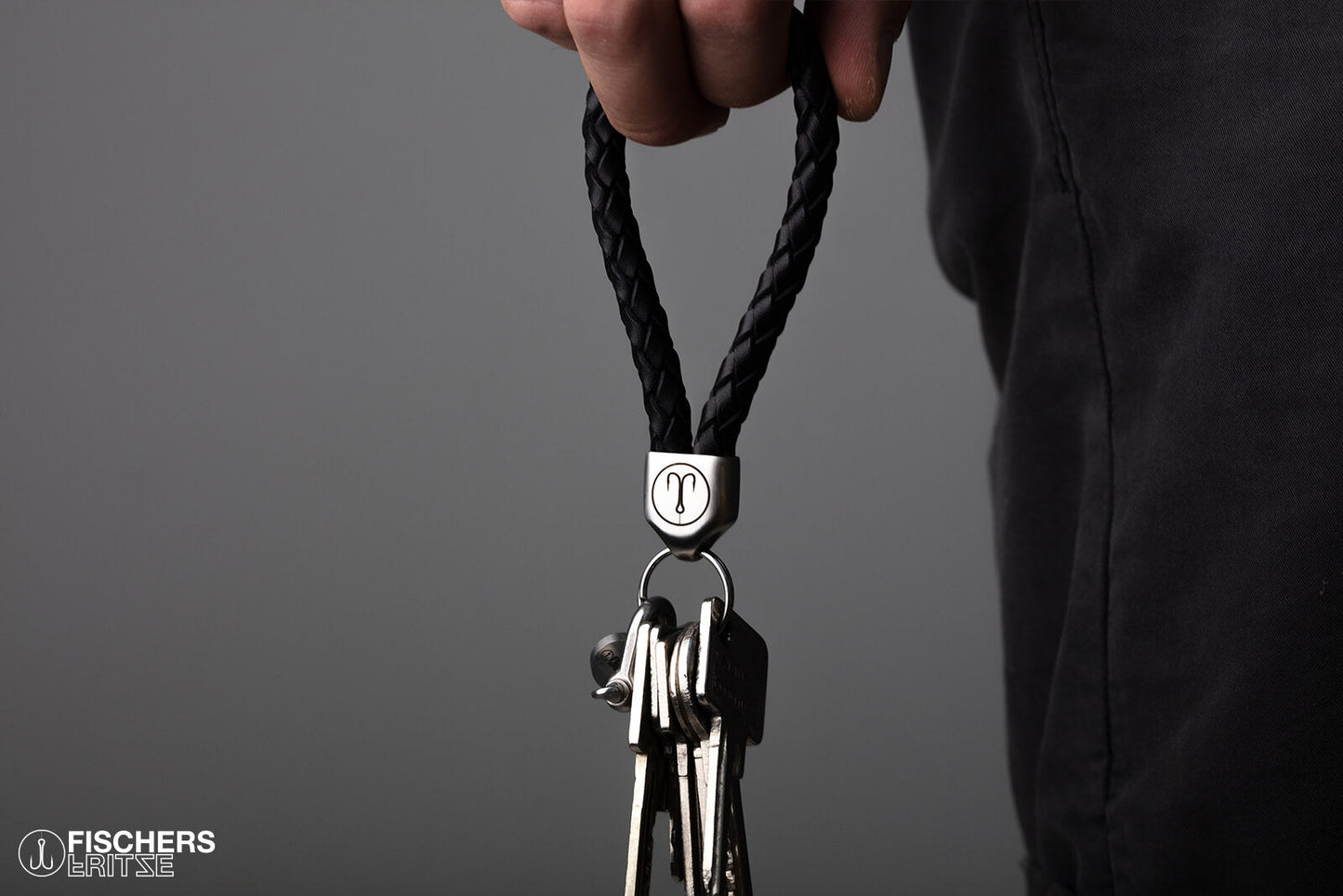 Hand haelt schwarzen Schluesselanhaenger Leder geflochten mit Schluesselbund, Edelstahlfassung mit Gravur sichtbar