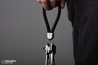 Hand haelt schwarzen Schluesselanhaenger Leder geflochten mit Schluesselbund, Edelstahlfassung mit Gravur sichtbar